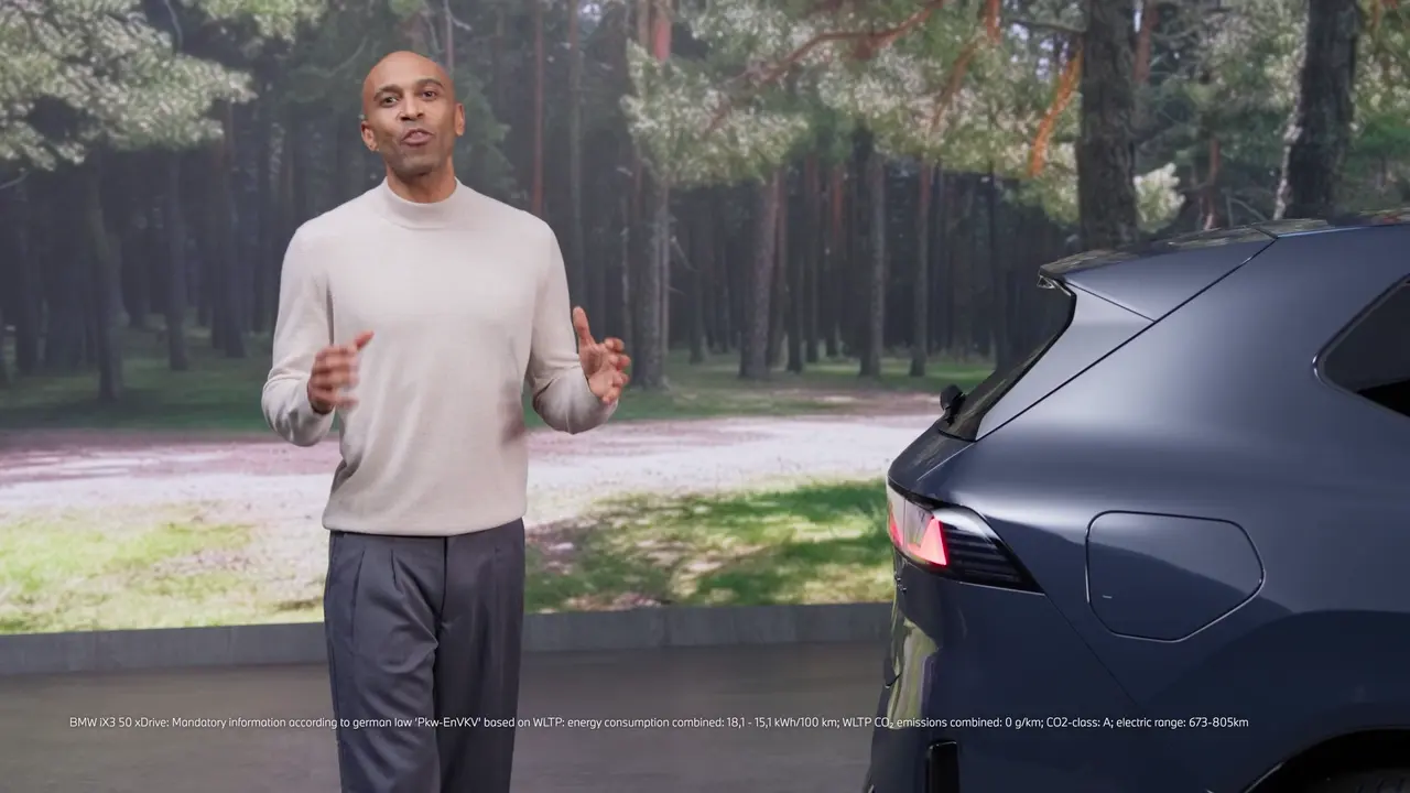 BMW presenter explaining vehicle-to-load beside the iX3 in a forest-themed studio